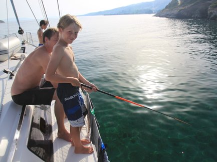 Ein Junge steht auf einem Boot und angelt, während ein Mann hinter ihm sitzt und die ruhige Wasseroberfläche betrachtet.