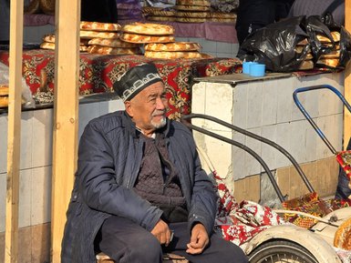 Ein älterer Mann sitzt entspannt auf einem Stuhl, umgeben von frischem Brot und einem geschäftigen Markt im Hintergrund.
