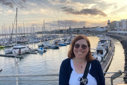 Eine lächelnde Frau mit Brille steht am Hafen, umgeben von vielen Booten und einem malerischen Sonnenuntergang im Hintergrund.