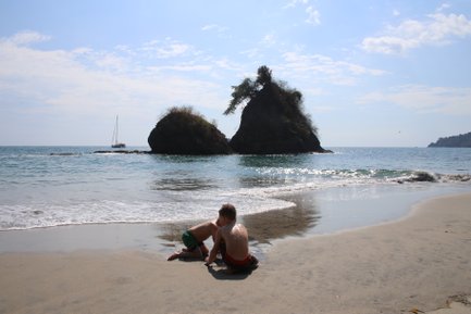 Zwei Kinder spielen am Strand, während sanfte Wellen gegen den Sand schlagen und Felsen im Hintergrund sichtbar sind.
