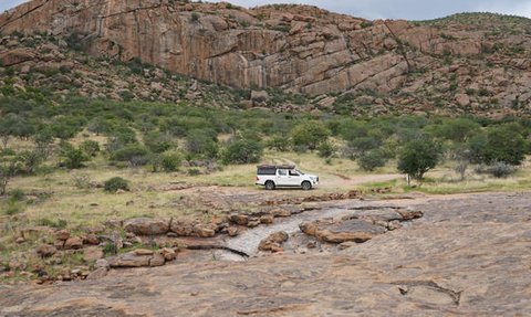Eine weiße Geländewagen steht auf einer felsigen Fläche, umgeben von grünen Bäumen und beeindruckenden Felsen im Hintergrund.