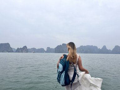 Eine Frau mit einem Rucksack steht am Wasser und blickt auf die beeindruckenden Felsen in der Ferne.