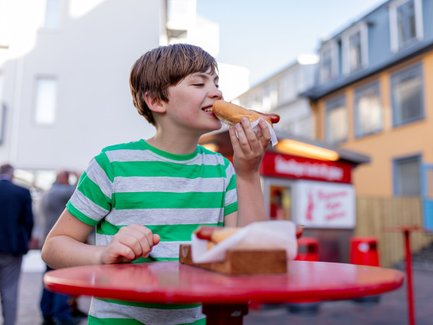 Ein Junge mit gestreiftem T-Shirt genießt einen Hotdog an einem roten Tisch in einer belebten Straße in Reykjavik.