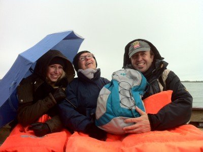 Drei Personen stehen unter einem blauen Regenschirm, lächeln und tragen orangefarbene Schwimmwesten auf einem Boot.