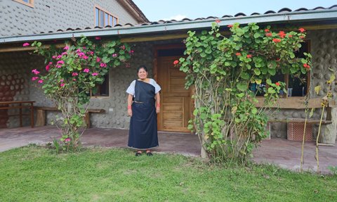 Eine Frau in traditioneller Kleidung steht vor einem Haus mit bunten Blumen und einem einladenden Eingang.
