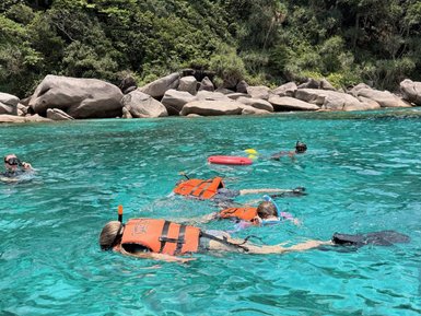 Kinder in orangefarbenen Schwimmwesten schnorcheln in klarem, türkisfarbenem Wasser vor einer bewaldeten Küste.