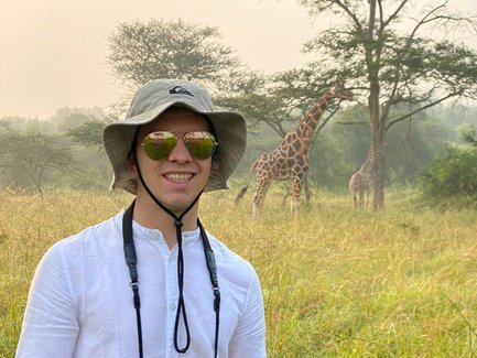 Ein junger Mann mit Sonnenbrille und Hut steht in einer grünen Savanne, während Giraffen im Hintergrund grasen.