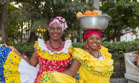Zwei fröhliche Frauen in bunten traditionellen Kleidern posieren in einem Park, eine trägt eine Schüssel mit Früchten auf dem Kopf.