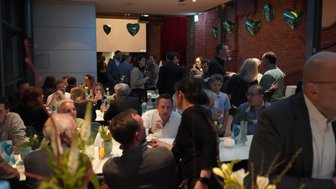 Eine lebhafte Veranstaltung mit Gästen, die an Tischen sitzen und Getränke genießen, umgeben von grünen Herzballons.