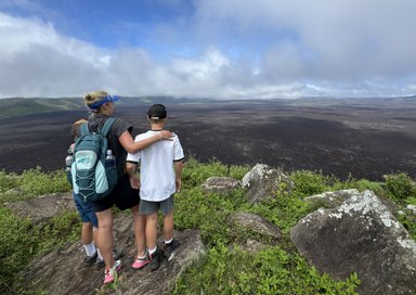 Eine Mutter steht mit zwei Kindern an einem Aussichtspunkt und blickt auf eine weite, vulkanische Landschaft.