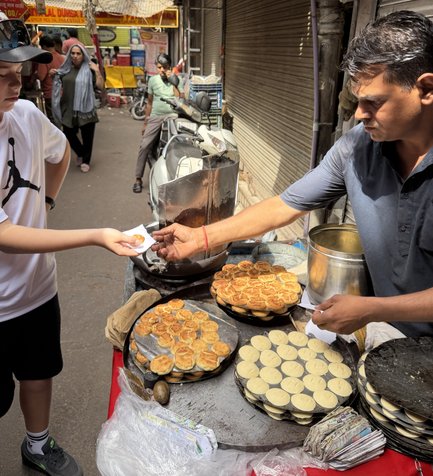 Ein Junge reicht einem Straßenverkäufer eine Bestellung, während köstliche Snacks auf einem Tisch in Delhi ausgestellt sind.