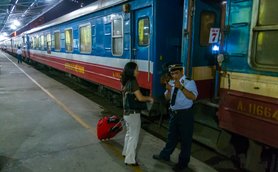 Eine Frau mit einem Koffer spricht mit einem Zugbegleiter an einem Bahnsteig in der Nacht.