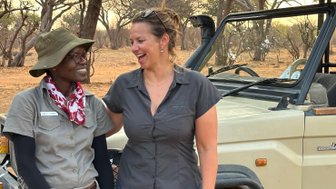 Zwei Frauen stehen lächelnd neben einem Jeep in der afrikanischen Savanne, umgeben von trockenen Bäumen und einem warmen Licht.