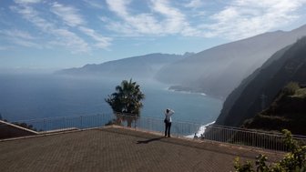 Eine Person steht auf einer Terrasse und blickt auf die atemberaubende Küstenlandschaft von Porto Moniz mit Bergen im Hintergrund.