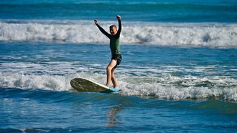Ein junger Surfer balanciert auf seinem Brett in den Wellen des Ozeans und zeigt seine Freude mit erhobenen Armen.