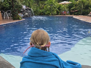 Ein Kind sitzt am Rand eines Pools, umhüllt von einem blauen Handtuch mit einem Elefantenmotiv, während es auf das Wasser blickt.