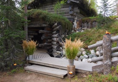 Ein rustikales Holzhaus mit einem begrünten Dach, umgeben von Bäumen und Steinen, bietet eine einladende Atmosphäre.