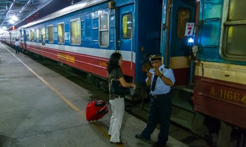 Eine Frau mit einem Koffer spricht mit einem Zugbegleiter an einem Bahnsteig in der Nacht.