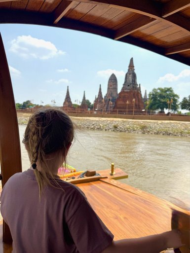 Ein Kind mit geflochtenem Haar blickt auf die beeindruckenden Tempelruinen von Ayutthaya, während es auf einem Boot sitzt.