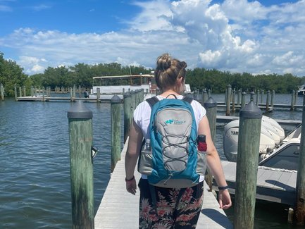 Eine Person mit einem Rucksack geht auf einem Steg entlang, umgeben von Wasser und Booten unter einem strahlend blauen Himmel.