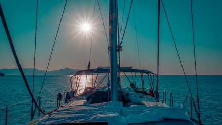 Ein Segelboot fährt über ruhiges Wasser, während die Sonne hell am Himmel strahlt und die Landschaft im Hintergrund sichtbar ist.