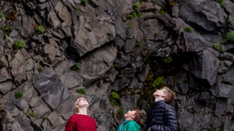Drei Kinder stehen auf einem Kiesstrand und schauen fasziniert nach oben zu den beeindruckenden Felsen.