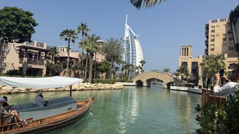 Ein traditionelles Boot fährt durch eine grüne Wasserstraße, umgeben von modernen und historischen Gebäuden in Dubai.