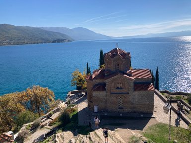 Eine alte Kirche mit roten Ziegeldächern steht majestätisch am Ufer des glitzernden Ohridsees, umgeben von Bäumen.