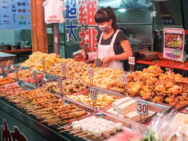 Eine Verkäuferin in einem Streetfood-Markt bereitet eine Vielzahl von frittierten Snacks und Meeresfrüchten vor.