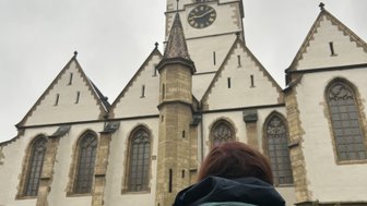 Eine Person mit einem Rucksack steht vor einer beeindruckenden Kirche mit einem hohen, spitzen Turm und großen Fenstern.