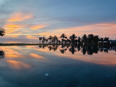 Ein atemberaubender Sonnenuntergang spiegelt sich im ruhigen Wasser eines Pools, umgeben von Palmen und einem friedlichen Himmel.