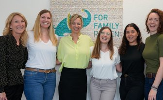 Sechs Frauen stehen lächelnd vor einer bunten Wand mit dem Schriftzug 'FOR FAMILY'. Sie wirken freundlich und einladend.