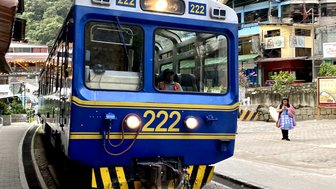 Ein blauer Zug mit der Nummer 222 steht an einem Bahnhof, umgeben von üppigen grünen Bergen und einem kleinen Dorf.