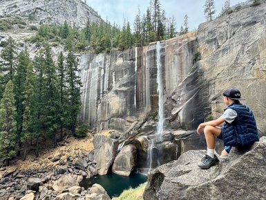 Ein Junge sitzt auf einem großen Felsen und blickt auf einen Wasserfall, umgeben von majestätischen Bäumen und Felsen.