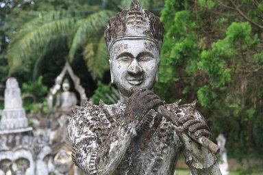 Eine alte Statue eines lächelnden Mannes, der eine Flöte spielt, umgeben von üppigem Grün und weiteren Skulpturen im Hintergrund.