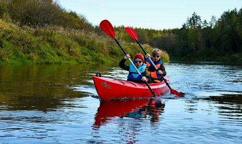 Zwei Kinder paddeln in einem roten Kajak auf einem ruhigen Fluss, umgeben von grünen Ufern und Bäumen im Hintergrund.