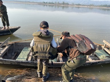 Ein Junge in einer Schwimmweste beobachtet einen Mann, der ein Boot am Ufer eines ruhigen Flusses vorbereitet.