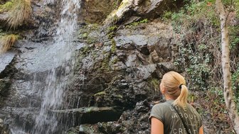 Eine Frau steht vor einem Wasserfall, der über eine felsige Klippe fließt, umgeben von üppigem Grün und Bäumen.