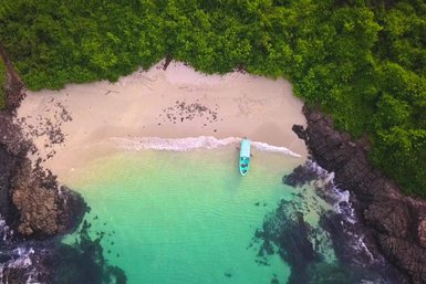 Ein malerischer Strand mit feinem Sand und klarem Wasser, umgeben von üppigem Grün und Felsen, bietet eine ruhige Atmosphäre.