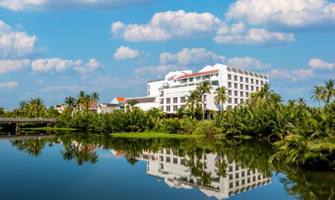 Ein modernes Hotelgebäude spiegelt sich im ruhigen Wasser eines Teiches, umgeben von üppigem Grün und Palmen.