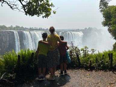 Eine Mutter umarmt ihre beiden Kinder, während sie die majestätischen Victoriafälle im Hintergrund bewundern.