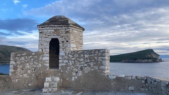 Eine alte Steinhütte mit einem gewölbten Dach steht auf einer Mauer, die über das Wasser und die umliegenden Hügel blickt.