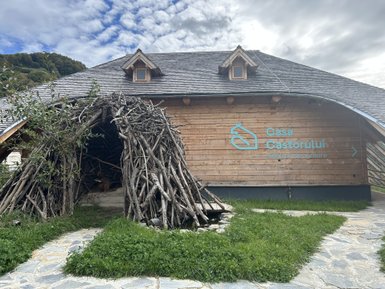 Ein Holzgebäude mit einem großen, kunstvoll gestalteten Eingang aus Ästen, umgeben von grünem Gras und einem gepflasterten Weg.