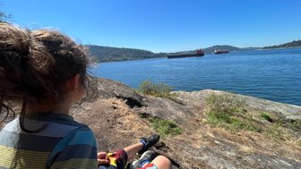 Ein Kind sitzt auf einem Felsen am Ufer eines ruhigen Sees und beobachtet zwei Schiffe, die sanft vorbeigleiten.