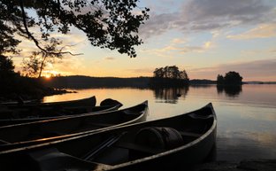 Am Ufer eines ruhigen Sees liegen mehrere Kanus, während die Sonne hinter den Bäumen untergeht und den Himmel erleuchtet.