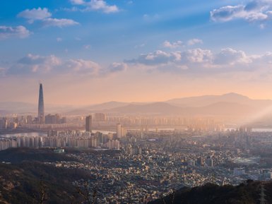 Die Skyline von Seoul bei Sonnenuntergang, mit dem markanten Lotte World Tower, umgeben von sanften Hügeln und Wolken.