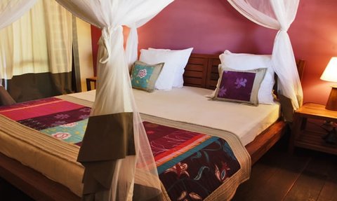 Ein gemütliches Schlafzimmer mit einem großen Bett, umgeben von einem weißen Moskitonetz und bunten Kissen auf der Bettdecke.