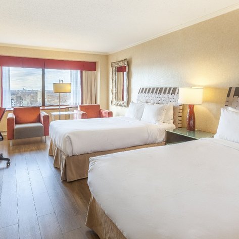 Ein modernes Hotelzimmer mit zwei großen Betten, einem Schreibtisch und einem Fenster, das einen Blick auf die Stadt bietet.
