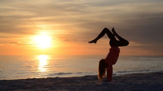 Ein Mädchen macht einen Handstand am Strand, während die Sonne im Hintergrund untergeht und den Himmel in warmen Farben färbt.