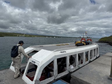 Ein Mann in beiger Kleidung nähert sich einem weißen Schnellboot am Ufer eines ruhigen Gewässers unter bewölktem Himmel.